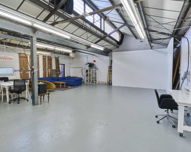 The image shows a spacious, well-lit studio with high ceilings, a large white backdrop, a seating area, and several tables and chairs arranged in the open space.