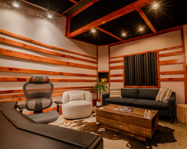 The image shows a cozy, modern lounge area featuring a dark sofa, a plush armchair, a wooden coffee table, and decorative wooden wall panels with recessed lighting.
