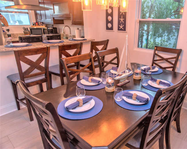 A dining table is set with blue and white plates, glasses, and decorative sailboat centerpieces, surrounded by wooden chairs in a well-lit room.