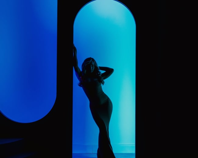 A silhouette of a woman in a fitted dress poses in a doorway illuminated by blue light.