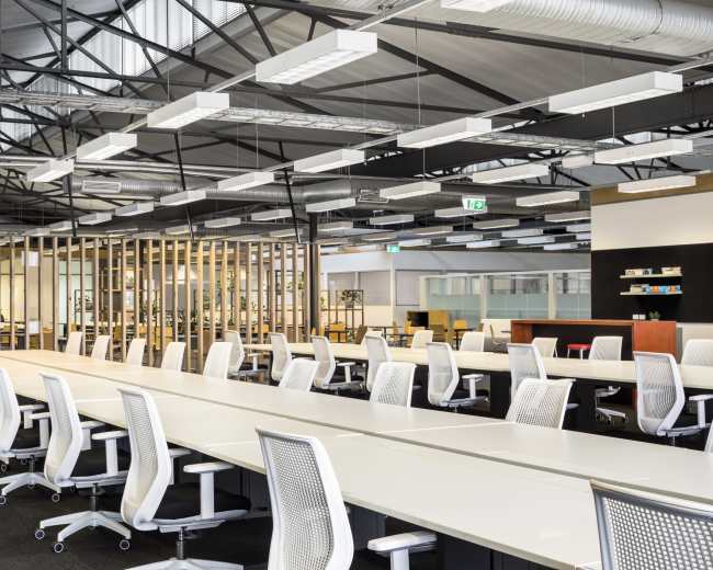 The image depicts a modern open-plan office space with long tables and ergonomic chairs arranged in neat rows under a high ceiling.