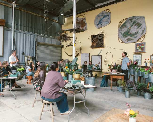 A group of people sits at tables in a workshop space filled with plants and artistic decorations while someone speaks at the front.