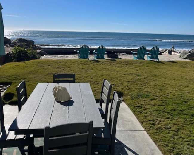 A wooden table and chairs are set on a grassy area overlooking the ocean and beach.