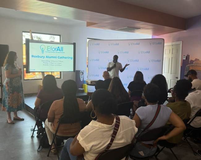 A speaker presents at the EforAll Roxbury Alumni Gathering in a well-lit room with an audience seated in rows.