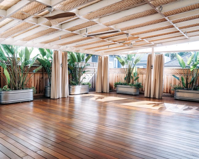 The image shows a spacious, covered wooden deck surrounded by tall green plants in large planters.