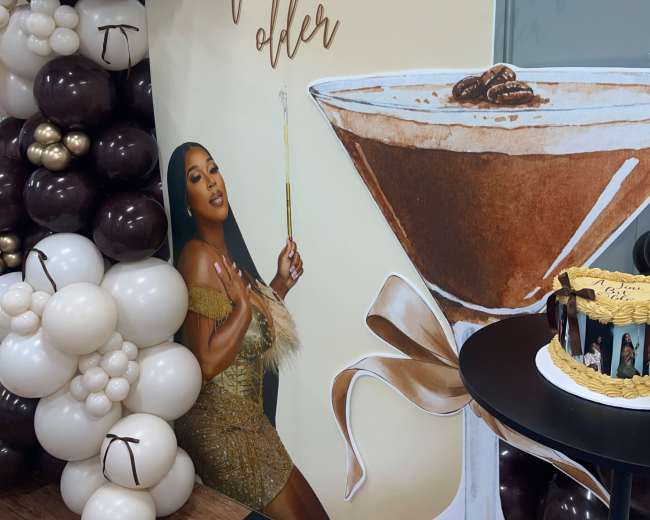 A large decorative display featuring a woman holding a cocktail is surrounded by balloons and a cake on a table.