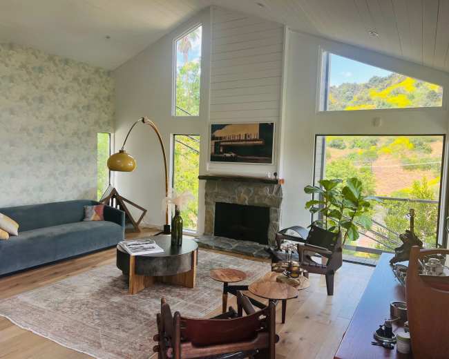 The image shows a modern living room with large windows, featuring a blue sofa, a stone fireplace, and a wooden table surrounded by chairs, all set against a backdrop of greenery outside.