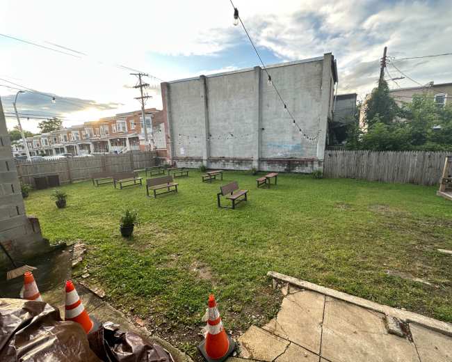 The image shows a grassy outdoor area with several wooden benches arranged in rows, surrounded by a wooden fence and a concrete walkway.
