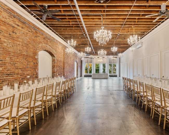 The image shows a spacious event hall with exposed brick walls, elegant chandeliers, and two long banquet tables arranged with white table settings.