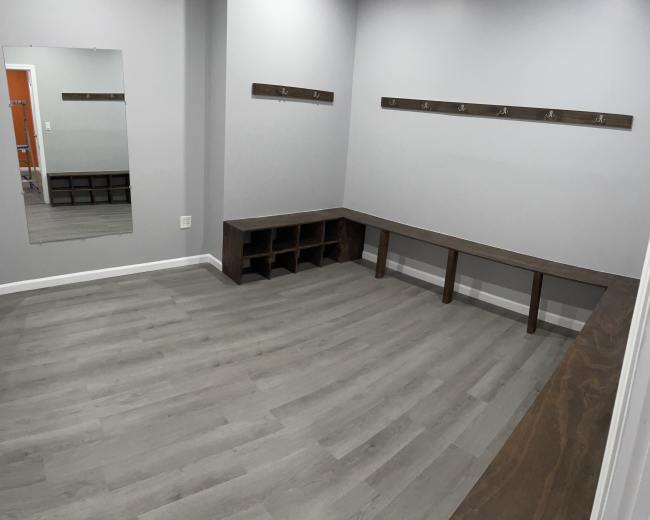 The image shows a minimalist changing room with a mirror, wooden bench, hooks on the wall, and a gray floor.
