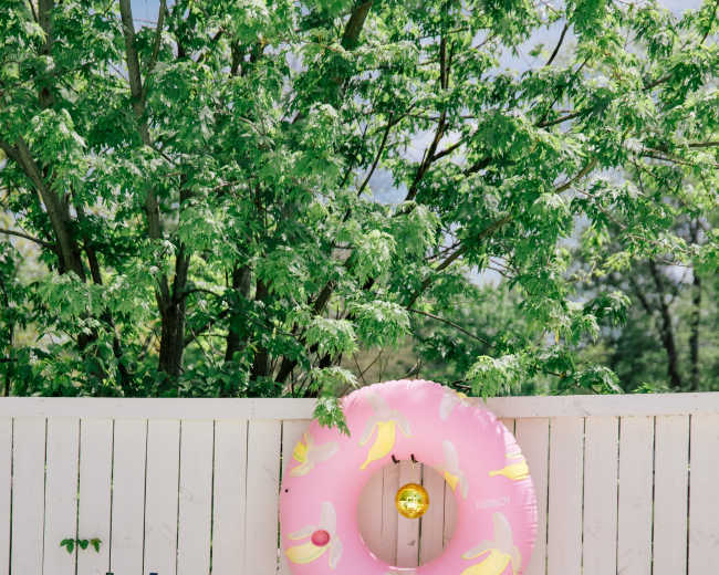 The scene features a pink inflatable donut float hanging on a white fence, with a bar cart and a cooler nearby amidst greenery.