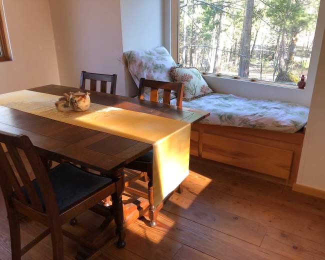 A wooden dining table with four chairs is set beside a window seat covered with a floral blanket and surrounded by trees outside.