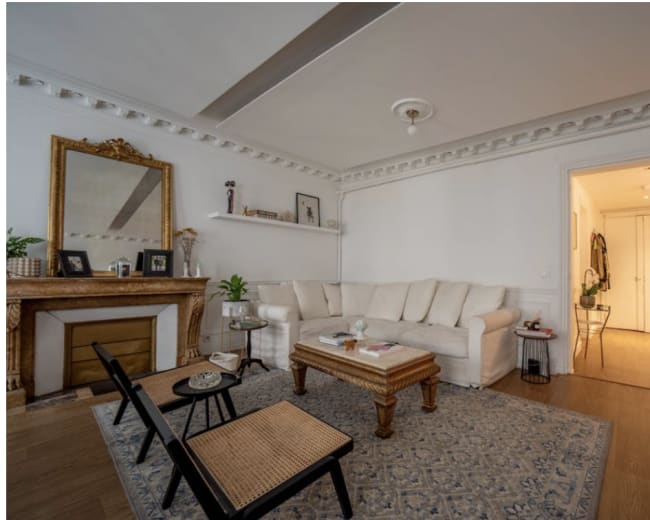 The image shows a bright living room with a beige sofa, a coffee table, and a decorative fireplace, which is connected to a hallway leading to other rooms.
