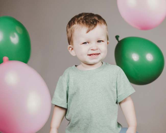 A smiling child stands in front of a gray background surrounded by colorful balloons.