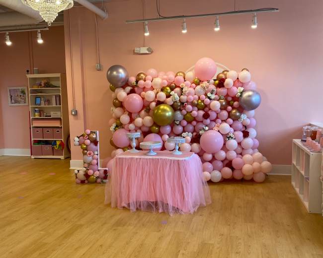 The image shows a decorated party area featuring a table with a white cloth, surrounded by a colorful balloon backdrop in shades of pink and gold.