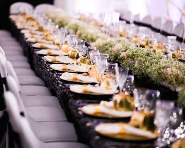 A long dining table is elegantly set with gold-accented plates, glasses, and decorative greenery down the center.