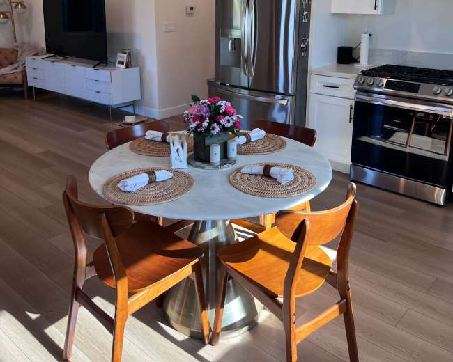 A modern kitchen features a round dining table set with placemats, four wooden chairs, and stainless steel appliances.