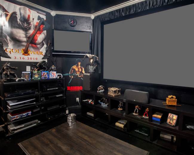 The image shows a home entertainment room with a large screen, gaming consoles, and various action figures on display, alongside themed posters on the walls.
