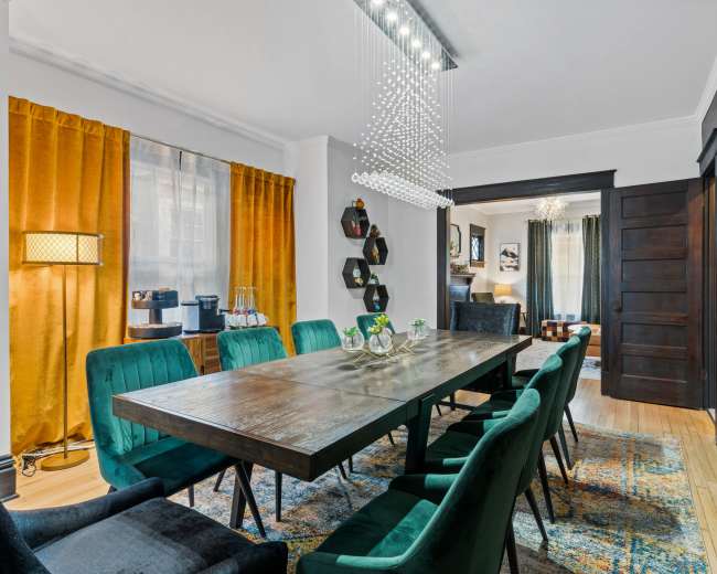 The image shows a dining room featuring a long wooden table surrounded by green upholstered chairs, with a modern chandelier overhead and yellow curtains framing a window.