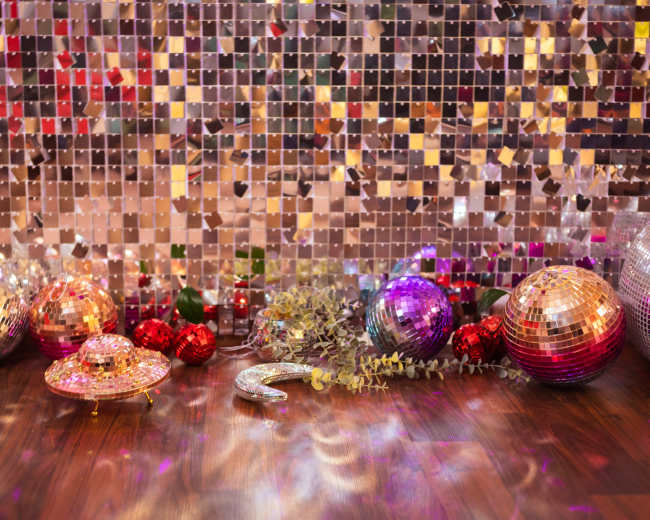 The scene features a sparkling backdrop of mirrored panels with various colorful disco balls, decorative ornaments, and greenery scattered on a wooden floor.