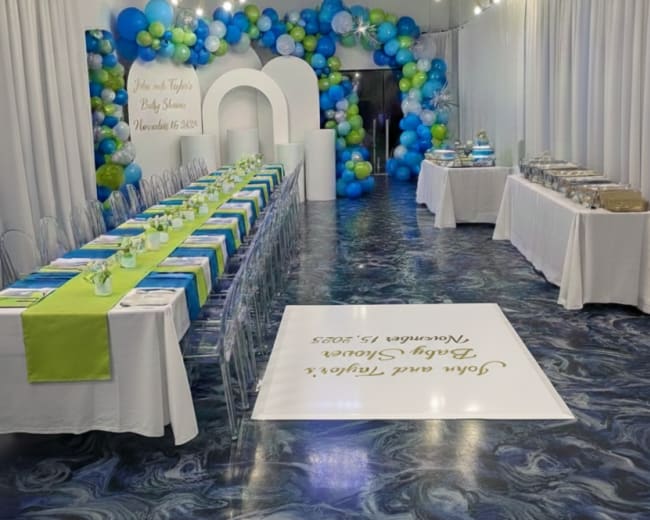 A decorated event space features a long table with blue and green table settings, colorful balloon garlands in the background, and a dessert display on the side.