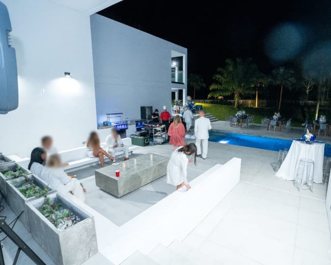 An outdoor gathering takes place at night by a swimming pool, with guests dressed in light clothing socializing in a modern, spacious environment.