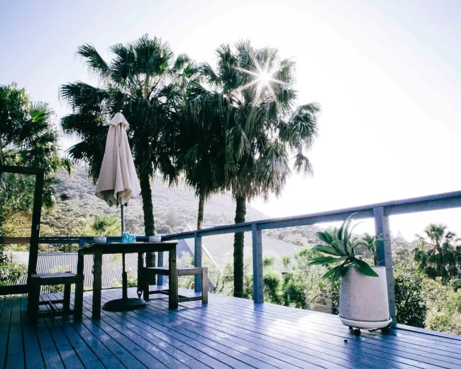 A wooden deck features a table and an umbrella, surrounded by palm trees and a view of nearby hills under a bright sun.