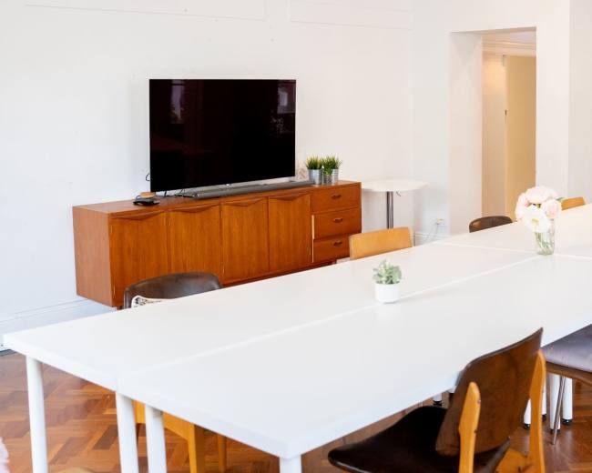 A modern meeting room features a long white table, wooden storage unit with a television on it, and light-colored decor.