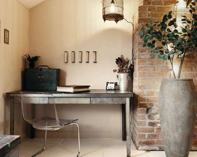 A small, minimalist workspace with a metal table, a transparent chair, and a brick wall adorned with decorative elements and a large vase.
