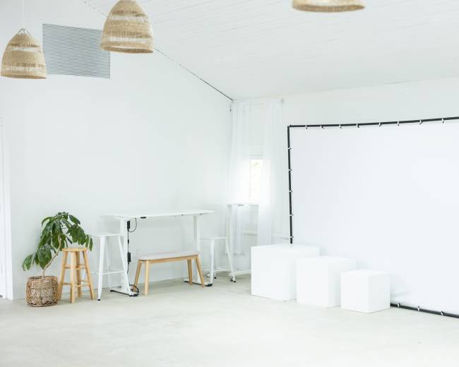 A bright, minimalistic room features a white backdrop setup, two white display cubes, a small leafy plant, and a wooden high table with stools.