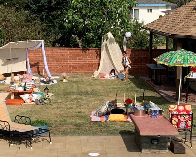The scene shows a backyard setting with a teepee structure, colorful blankets, and various decorations scattered on the grass near a pool and a shaded patio area.