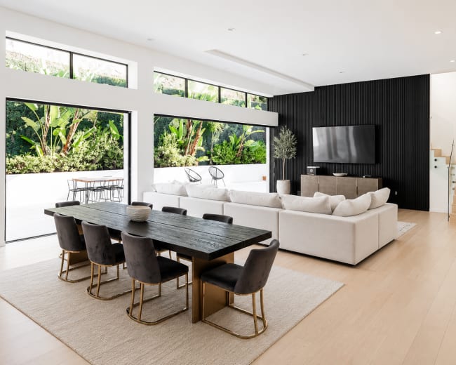 The image shows a modern open-concept living area featuring a dining table with eight chairs, a large sectional sofa, a television mounted on a black wall, and large windows overlooking greenery.
