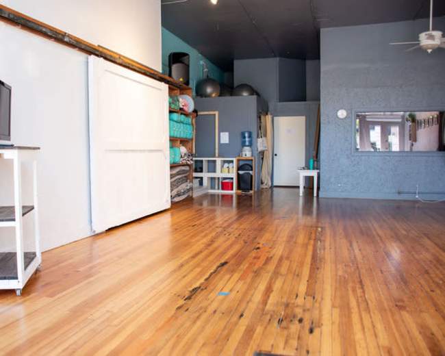 A spacious room with hardwood floors, featuring a television on a cart, storage shelves, and a small table.