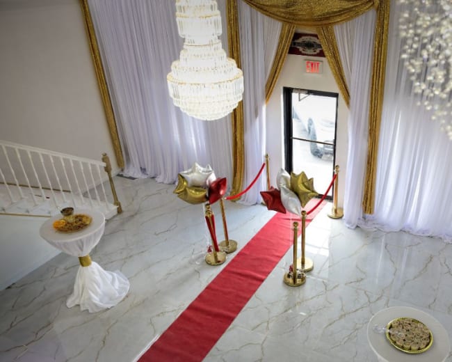 The image shows a grand entrance with a red carpet, flanked by gold and silver balloons, leading to an exit adorned with sheer white curtains and elegant chandeliers overhead.