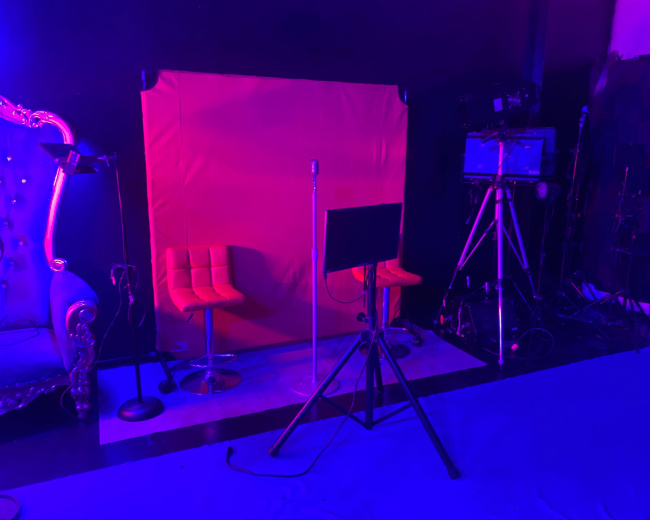 A studio setup with two orange chairs, a red backdrop, a microphone stand, and various lighting and filming equipment.