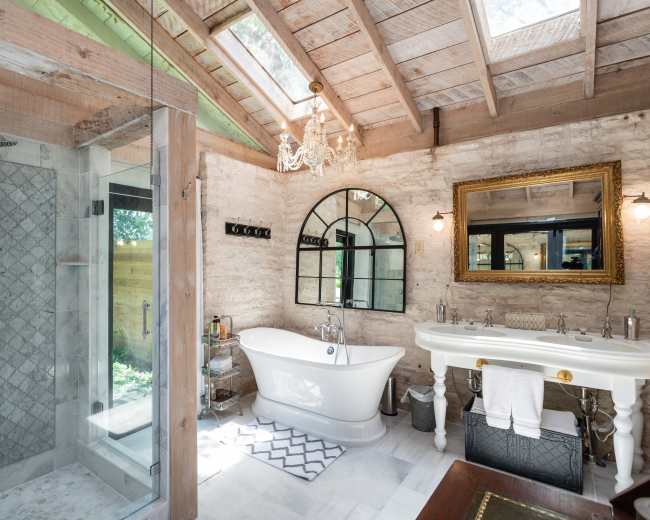 The bathroom features a freestanding tub, a glass shower enclosure, and a large mirror with natural light streaming in from the skylights.