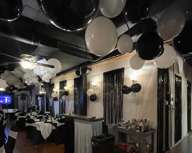 The image shows a decorated restaurant interior with black and white balloons hanging from the ceiling, tables set with white tablecloths, and a bar area stocked with beverages.