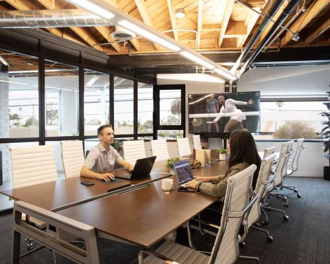A modern conference room features a long wooden table, a laptop-wielding man, a woman using a laptop, and a large screen displaying a presentation.