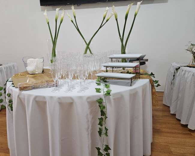 A round table covered with a tablecloth holds glasses, a tiered serving tray, and decorative elements, surrounded by two smaller tables adorned with white tablecloths and floral arrangements.
