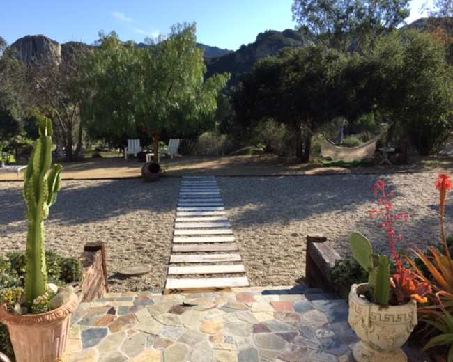A gravel path with wooden planks leads through a landscaped garden featuring cacti and a hammock under trees, with mountains in the background.