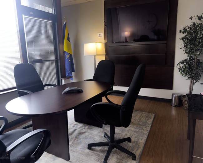 The image shows an empty conference room with a rectangular table and four black chairs, featuring a television mounted on the wall and a plant in the corner.
