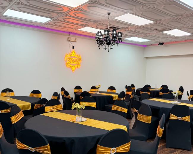 A banquet hall is set up with round tables covered in black cloth and adorned with gold accents, featuring centerpieces of flowers.
