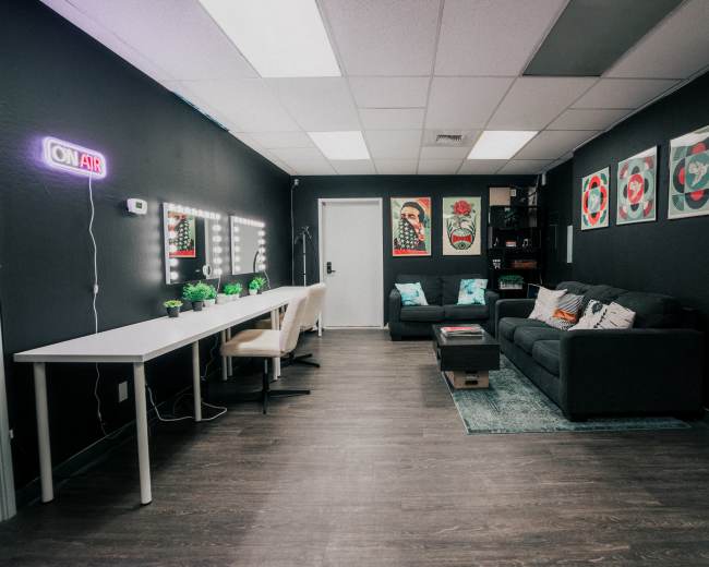A modern office space features a gray sofa, a white desk with chairs, and colorful wall art, illuminated by a neon "ON AIR" sign.