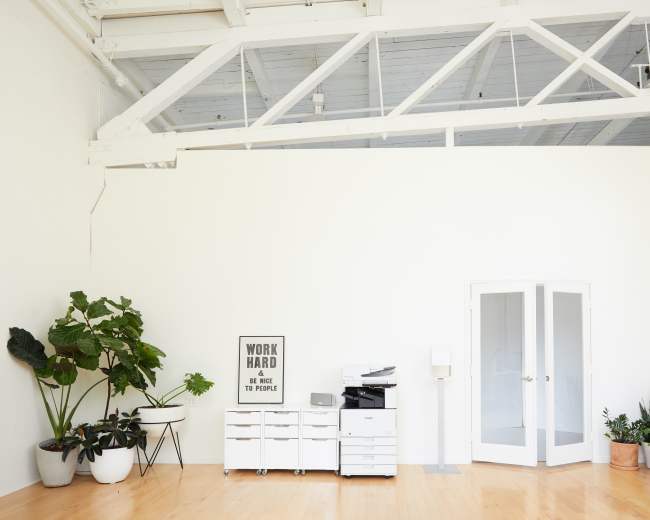 The image shows a minimalist office space featuring a printer stand, a glass door, large potted plants, and a motivational sign on the wall.