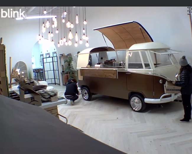 Two people work on a vintage-style food truck inside a well-lit, modern interior space with wooden flooring and hanging light fixtures.