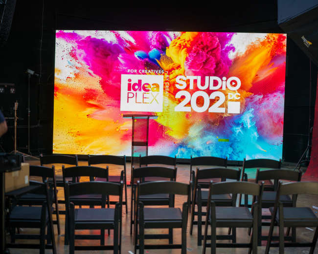 A brightly colored digital backdrop displays the words "idea PLEX" and "STUDIO 2021" in an empty auditorium set with black chairs.