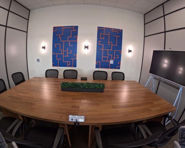 The image shows a conference room with a large wooden table surrounded by black chairs, two abstract blue and orange artworks on the walls, and a digital screen on a stand.