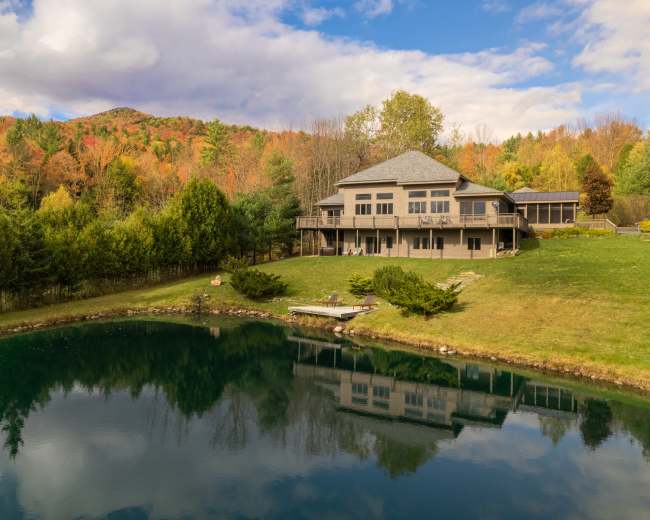 The image shows a large house with a deck overlooking a pond, surrounded by trees with autumn foliage and a mountainous background.