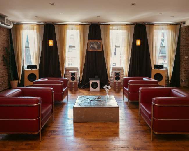 The image shows a modern lounge area with red armchairs arranged around a low, rectangular coffee table, set against a backdrop of large windows covered with flowing curtains.