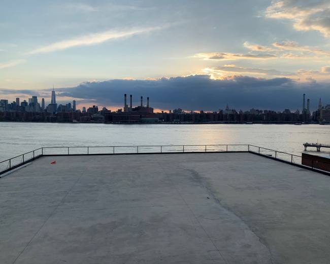 The image shows a waterfront view of a city skyline at sunset with a clear expanse of water in the foreground.